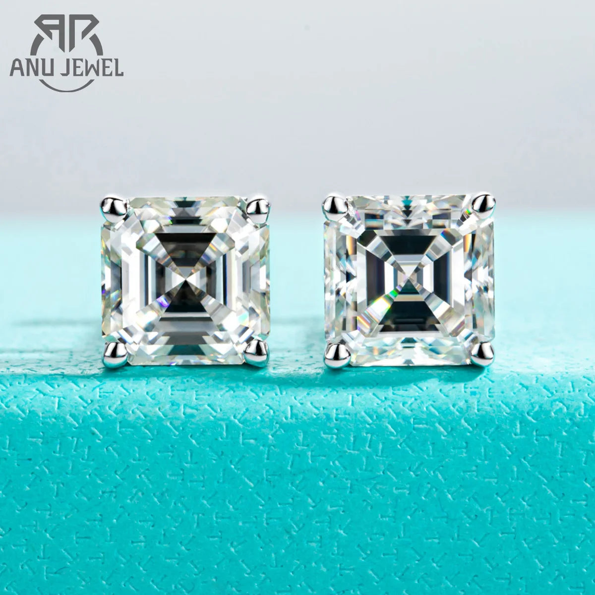 Pair of square-cut diamond earrings on a turquoise surface with 'ANU JEWEL' branding.