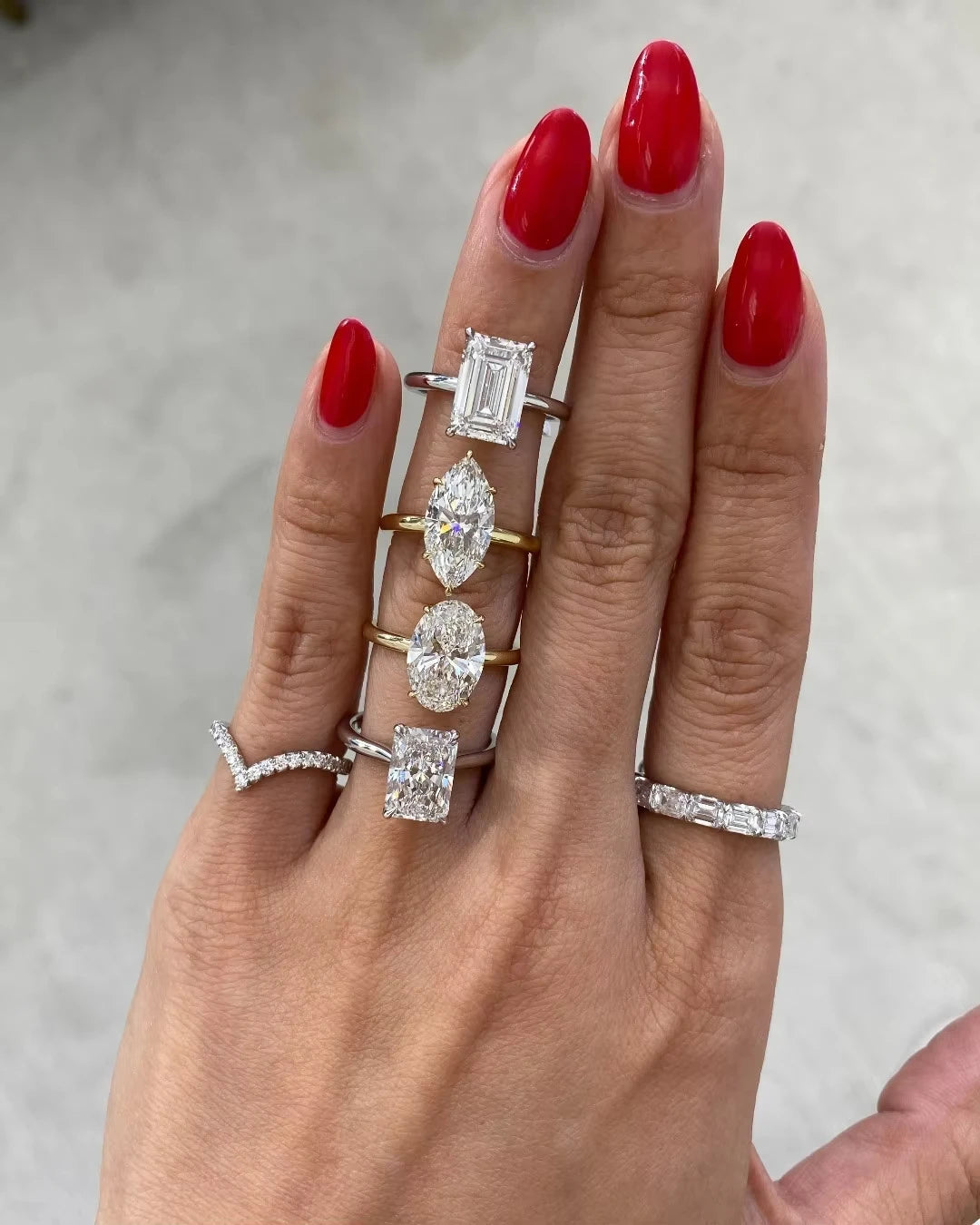 Hand wearing multiple diamond rings with red nail polish on a neutral background